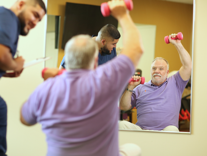 male senior fitness instructor works with male park place resident lifting weights in front of mirror