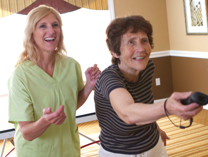 female resident aide assists senior female resident with wii bowling