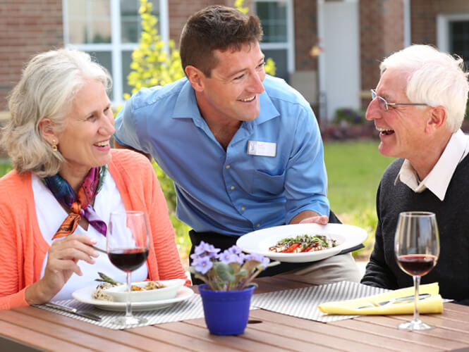 an older adult couple eating outdoors and enjoying victorian village scenery and being served by a man