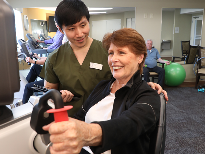 an elderly woman riding an exercise bike as male cna assists