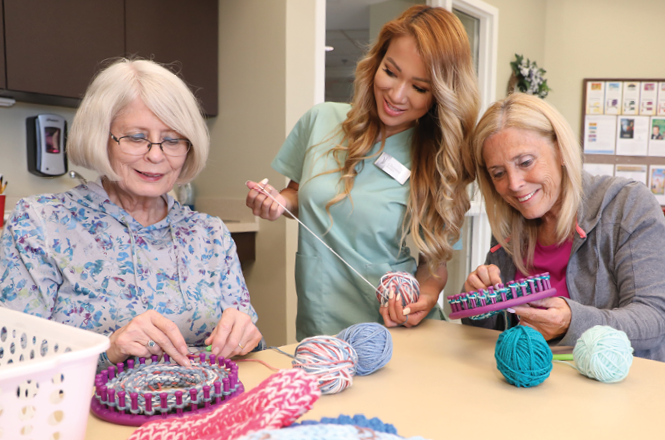 female CNA assists two female resident make hat on loom