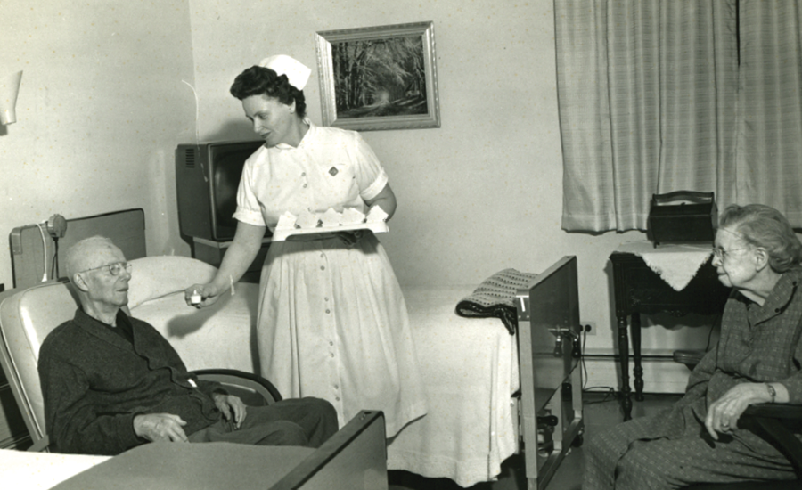 a nurse from the 1960s gives medicine to a senior male resident as his wife looks on