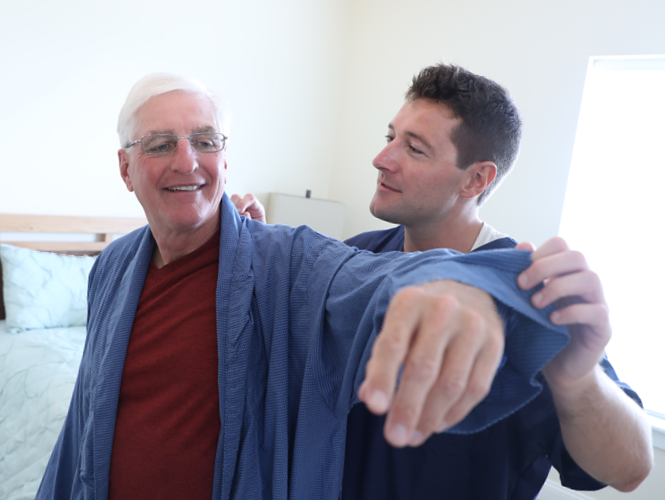 a male CNA assists male senior transition to home resident with robe